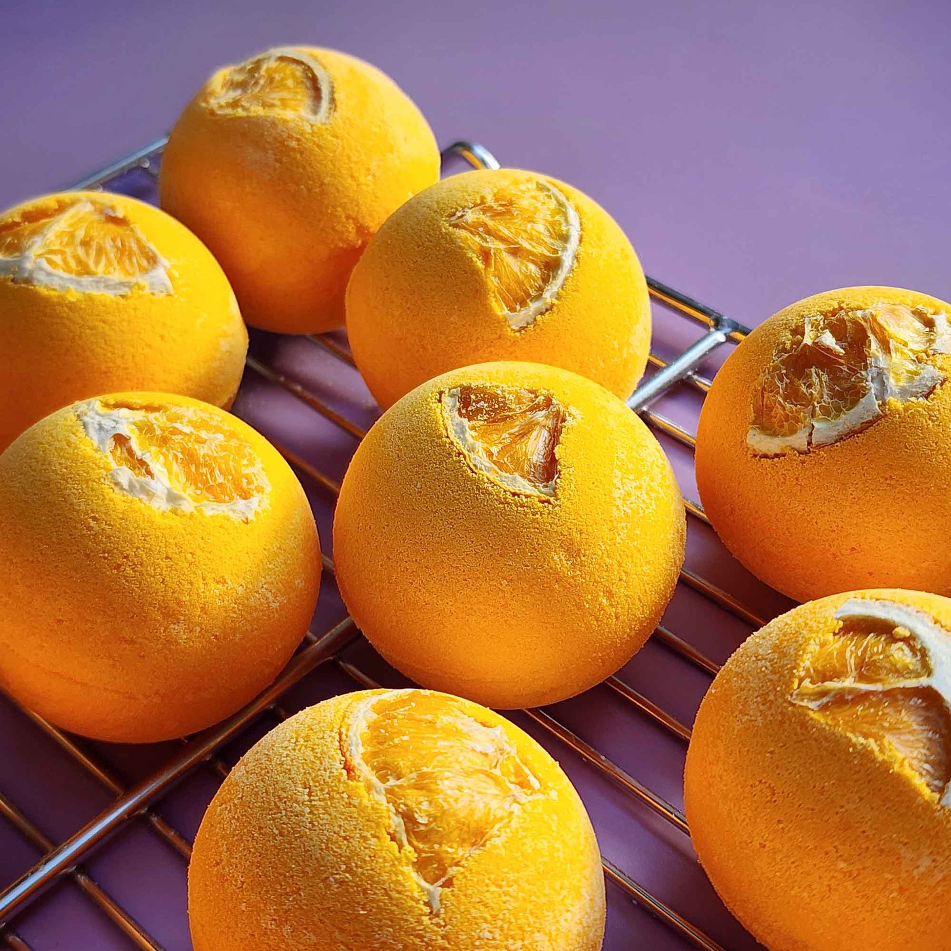 Bright yellow Energise aromatherapy bath bombs topped with dried orange slices cooling on a metal rack over a purple surface
