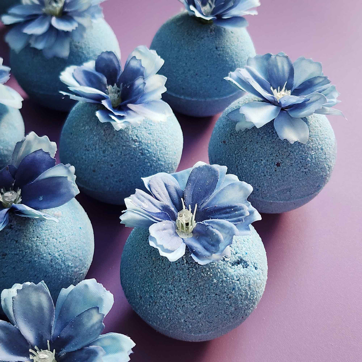 Close-up of handmade wildflower-scented bath bombs in dusty blue tones, each topped with blue faux petals on mauve background