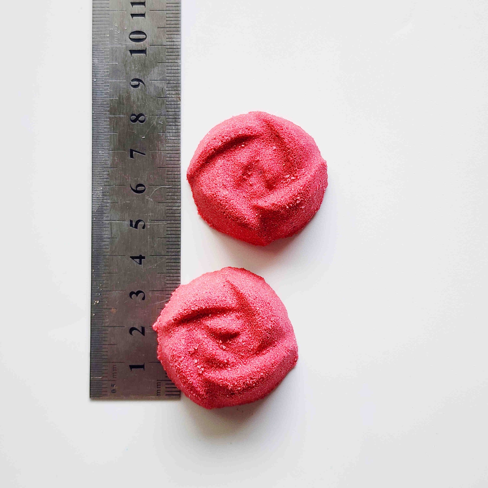rose shaped mini bath bombs coloured a deep red laid out next to a metal ruler on a white background