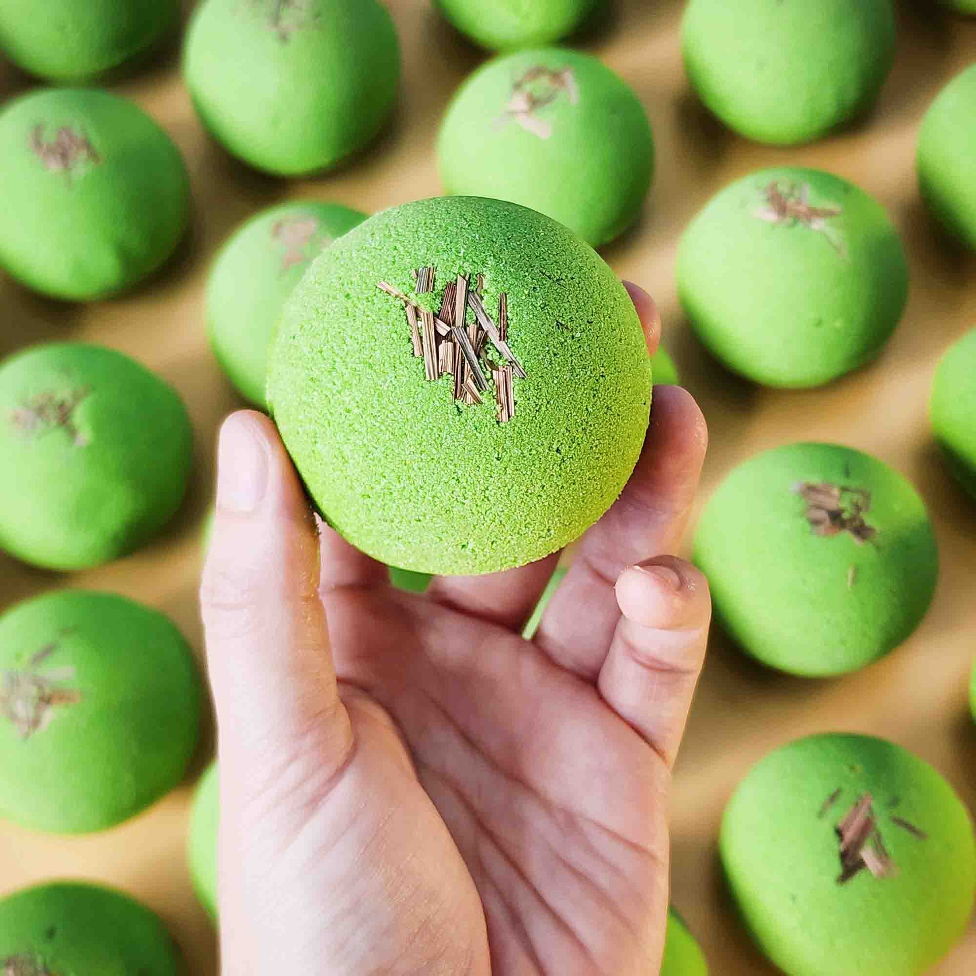 A hand holding a green Repair aromatherapy bath bomb with dried lemongrass, surrounded by more bath bombs in the background