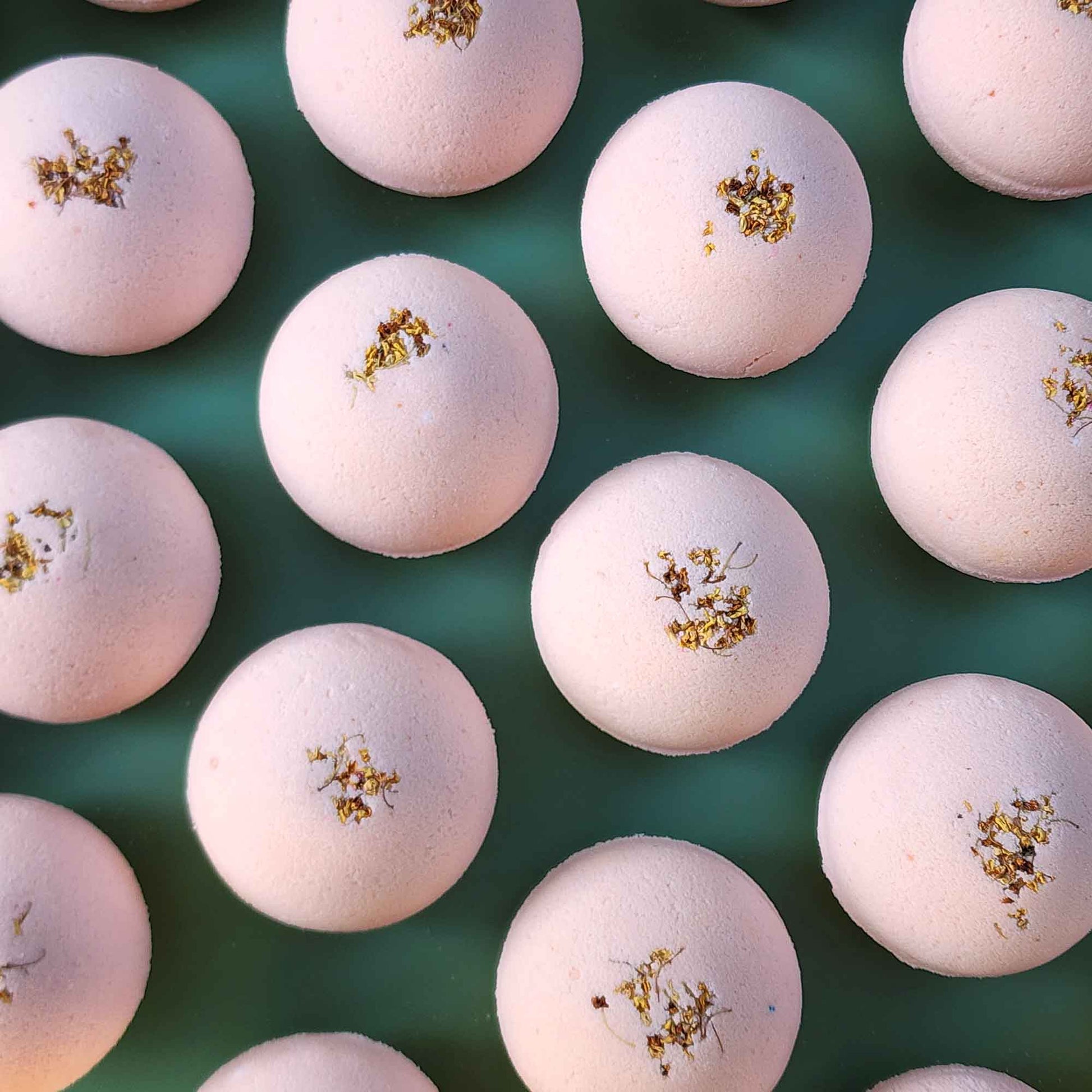Multiple pale pink Grounding aromatherapy bath bombs topped with small dried osmanthus flowers, arranged on a soft green back
