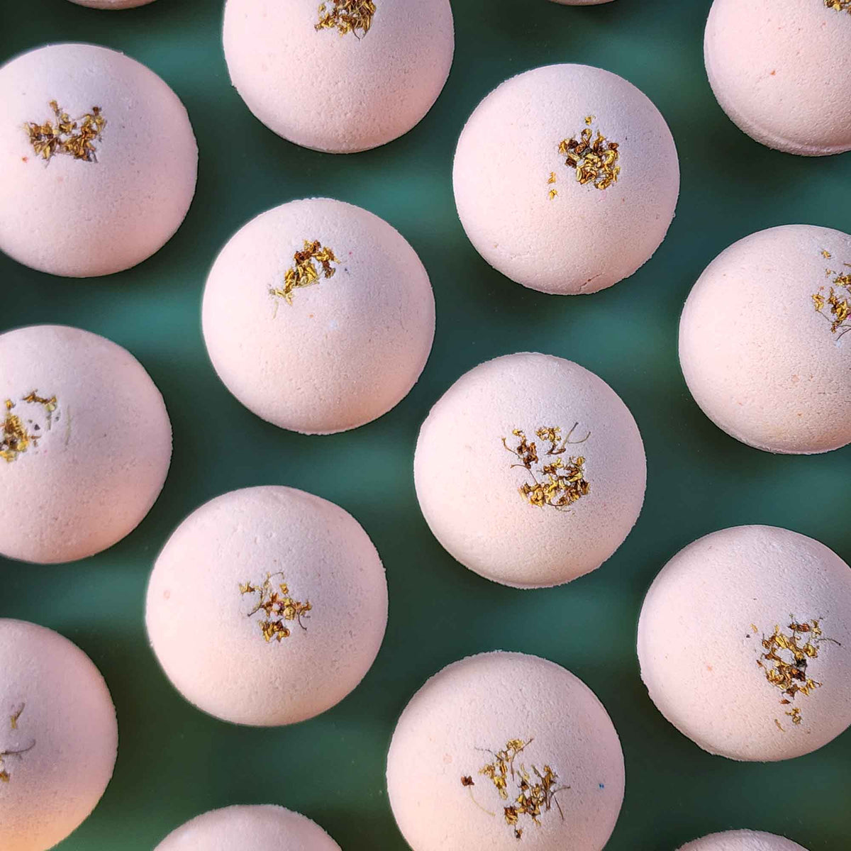 Multiple pale pink Grounding aromatherapy bath bombs topped with small dried osmanthus flowers, arranged on a soft green back