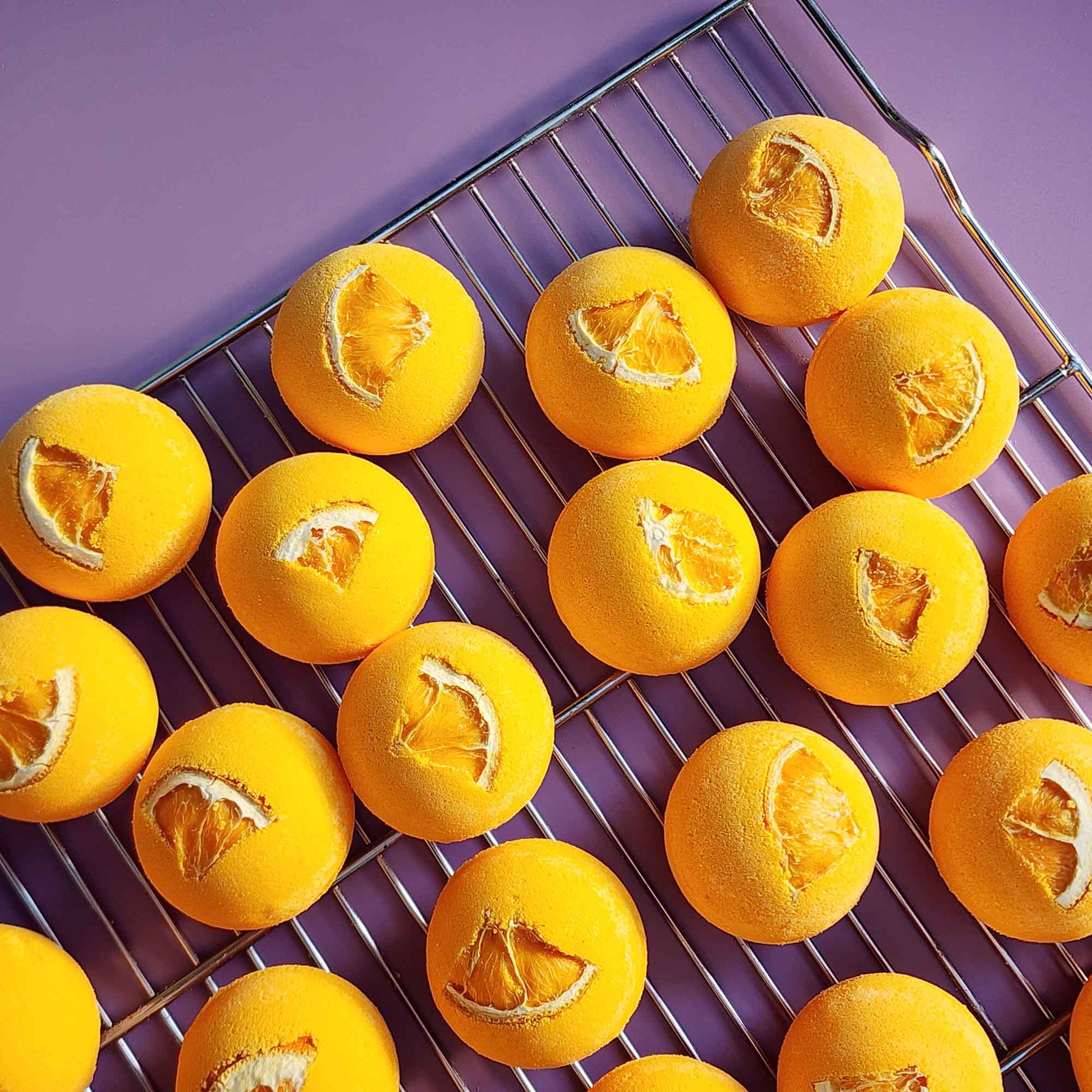 Bright yellow Energise aromatherapy bath bombs topped with dried orange slices cooling on a metal rack over a purple surface