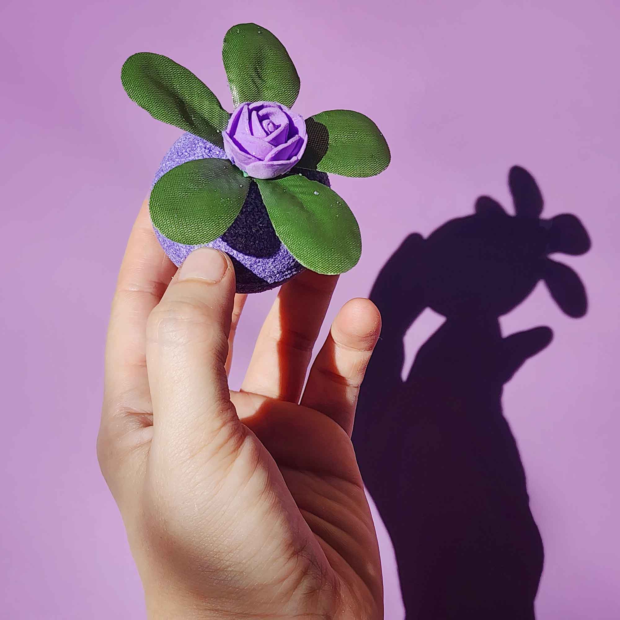 Hand holding a purple Wild Berry bath bomb with green leaves and a violet rose