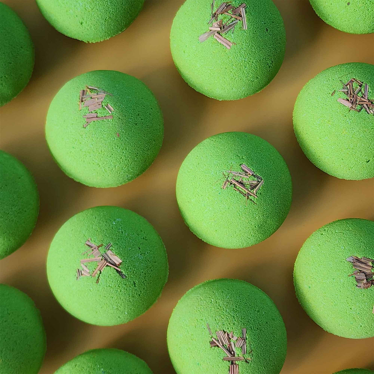 Multiple bright green Repair aromatherapy bath bombs topped with small pieces of dried lemongrass on a warm yellow background