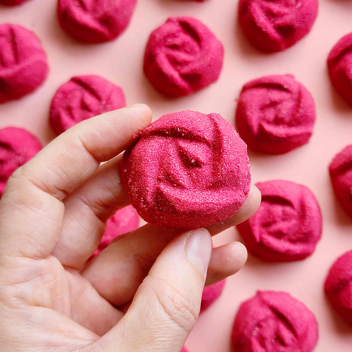 rose shaped mini bath bombs coloured a deep red laid out neatly on a surface with one held in a hand in the foreground