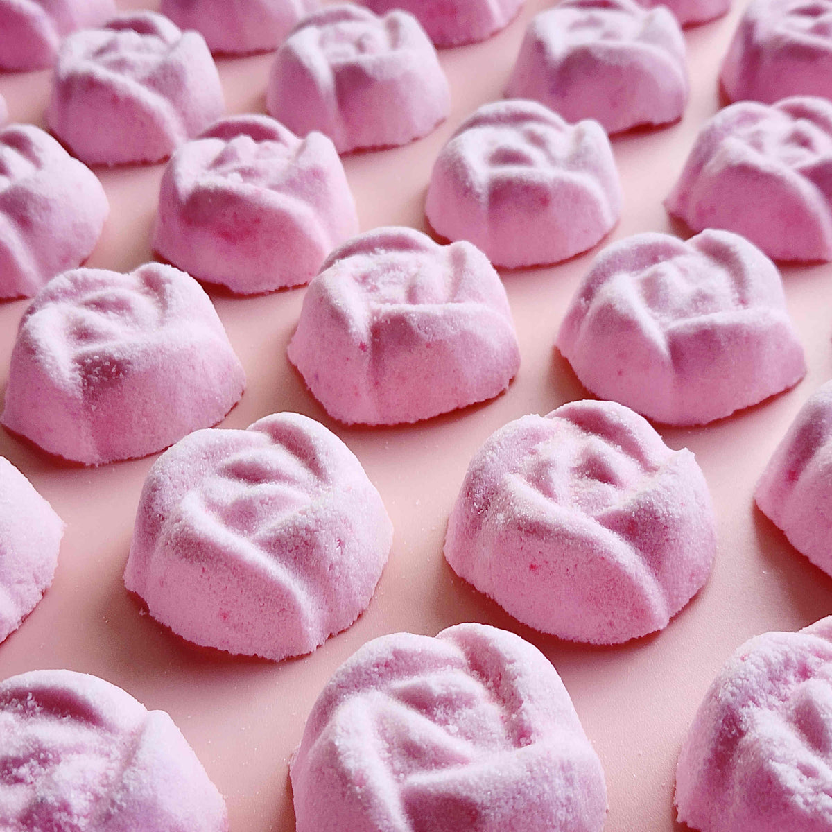 light dusty pink coloured mini bath bombs in the shape of roses laid out neatly on a plain pink background