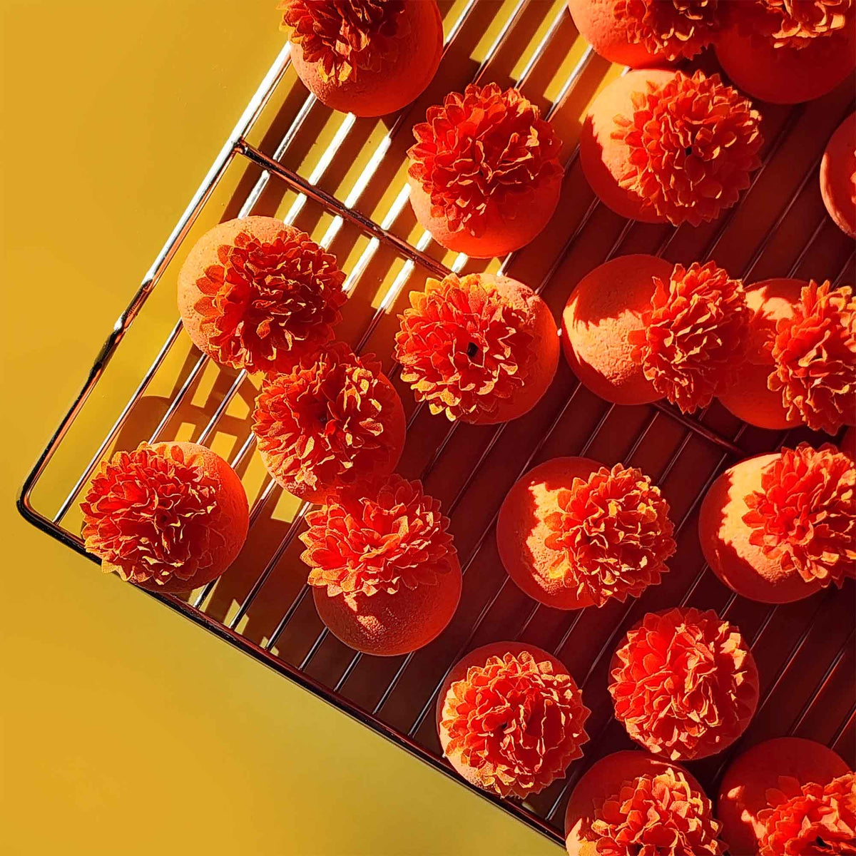 Bright orange bath bombs topped with faux calendula flowers drying on a metal rack under warm sunlight.