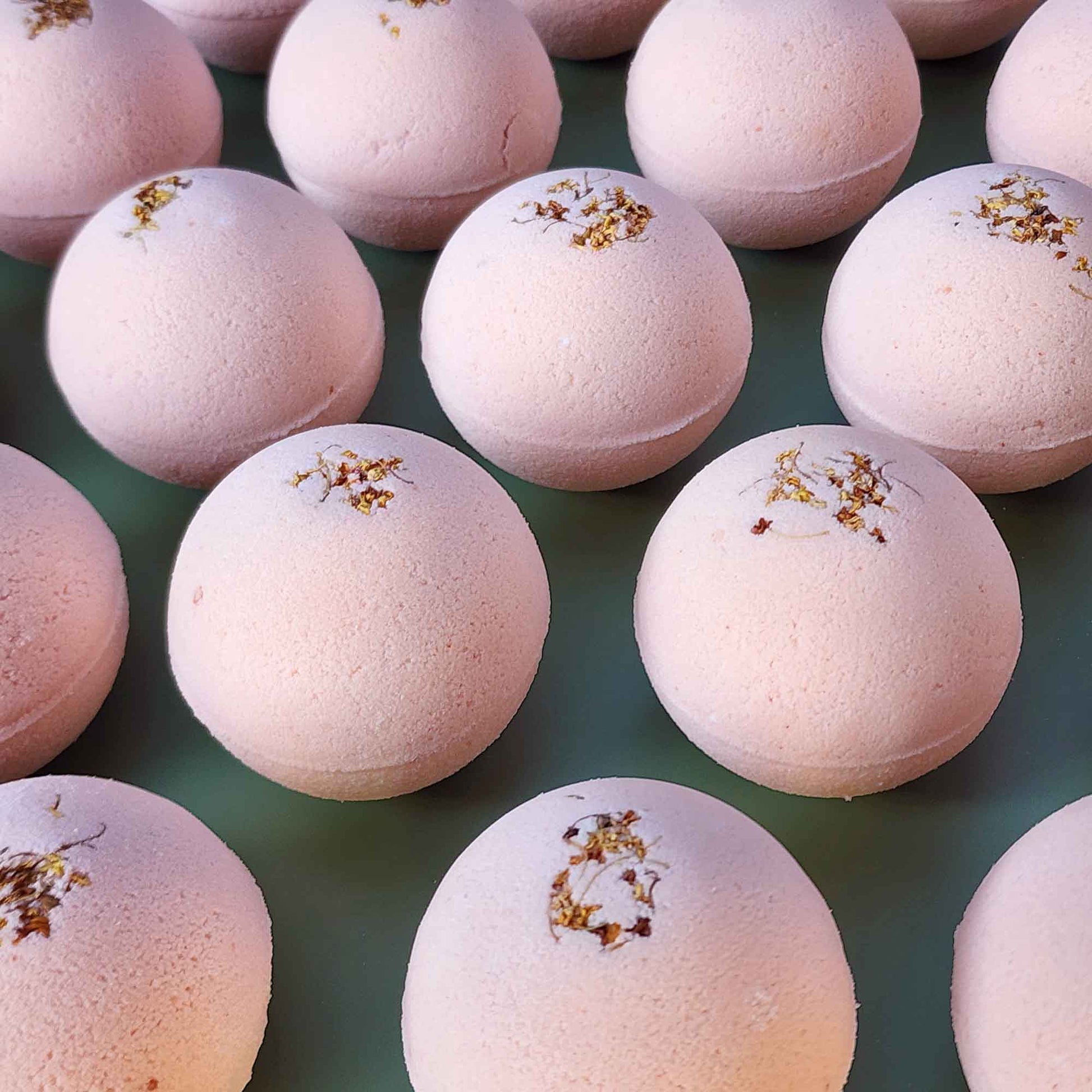 Multiple pale pink Grounding aromatherapy bath bombs topped with small dried osmanthus flowers, arranged on a soft green back