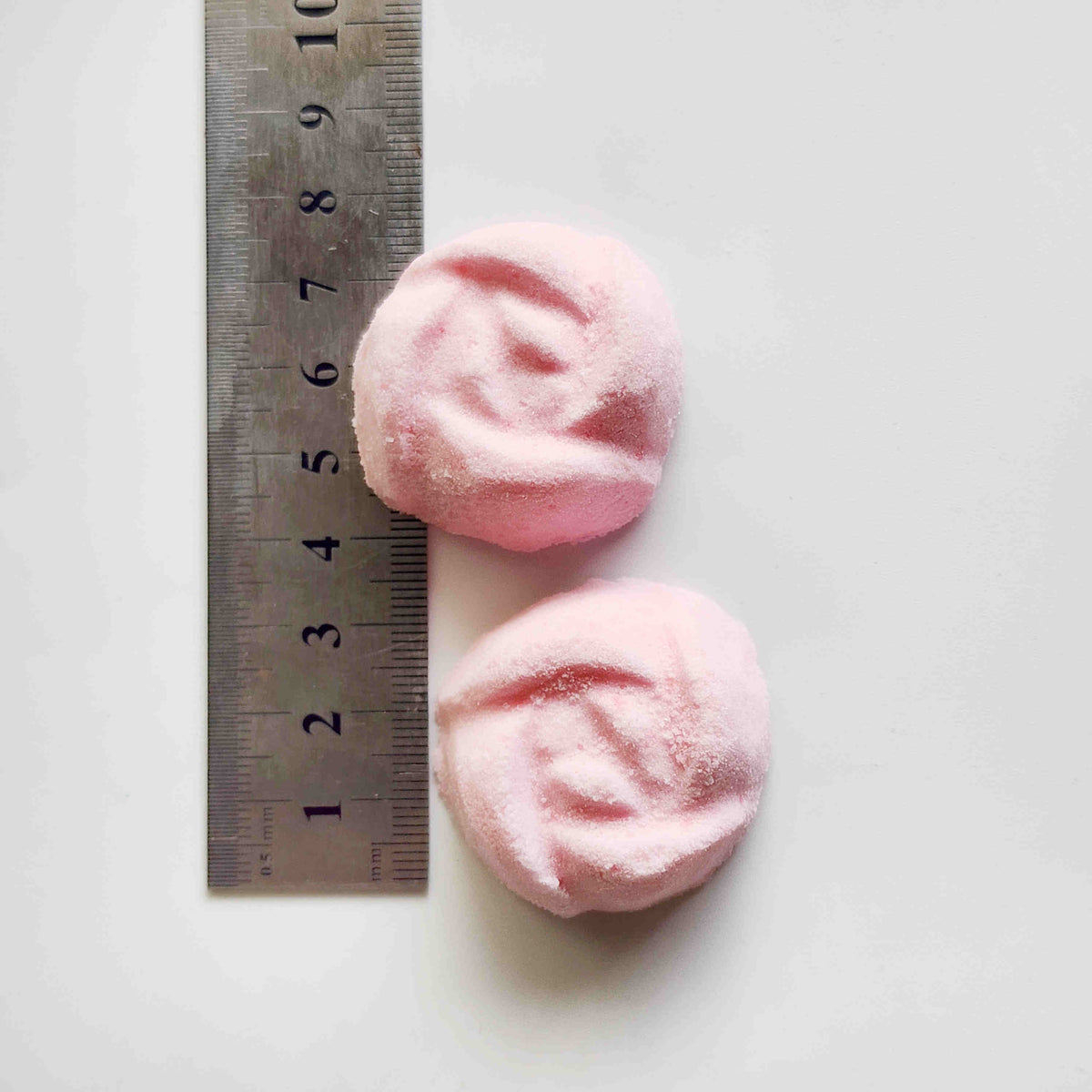 light dusty pink coloured mini bath bombs in the shape of roses placed next to a metal ruler