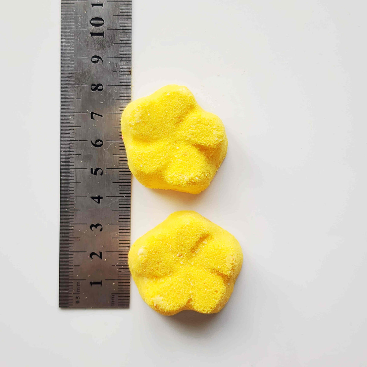 yellow frangipani shaped bath bombs placed next to a metal ruler on a white background