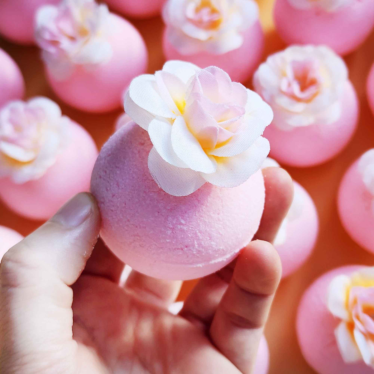 Hand holding a pink magnolia bath bomb topped with a soft faux flower, with others blurred behind