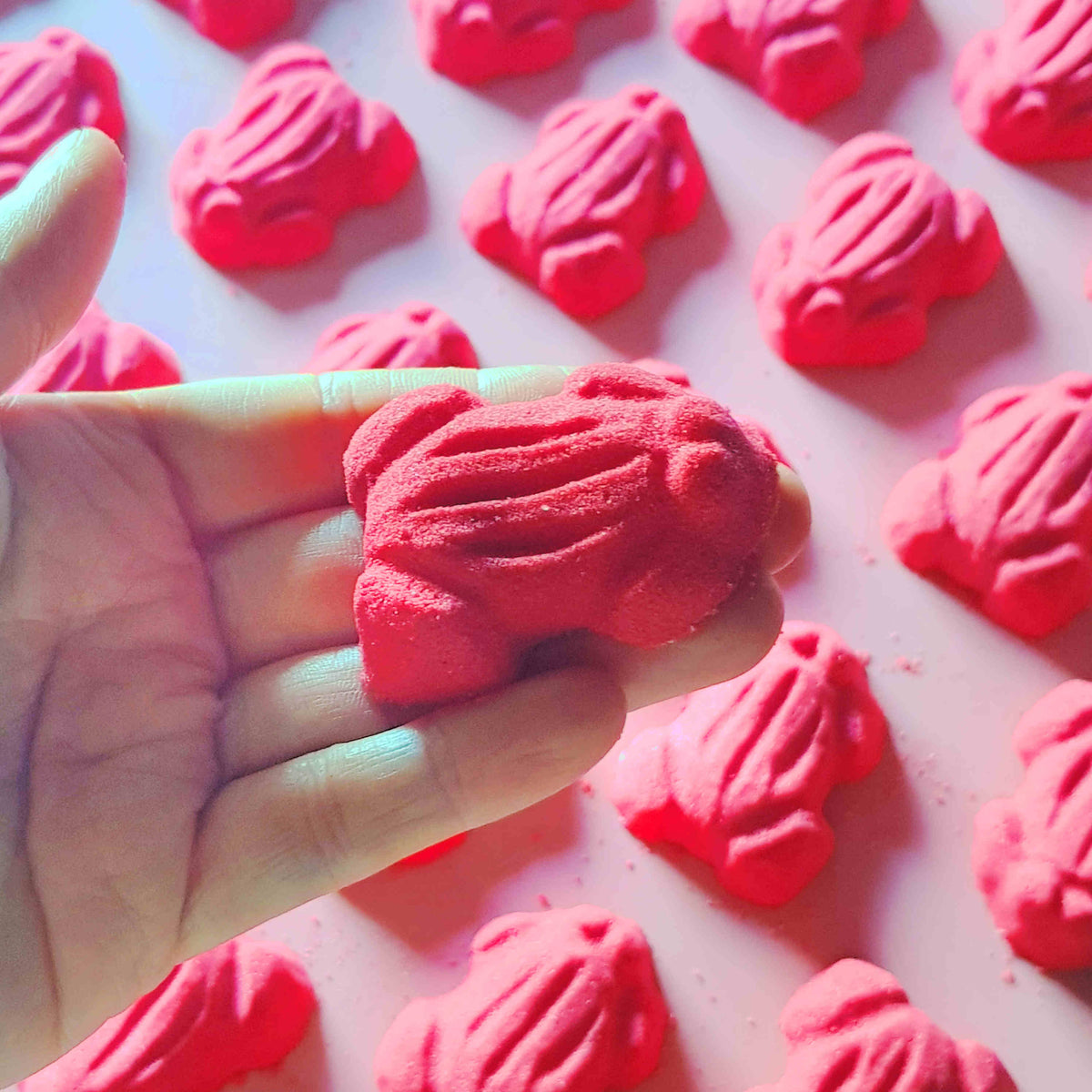 Red frog gummy candy shaped bath bombs layed out in an array over a plain red background with one being held in a hand